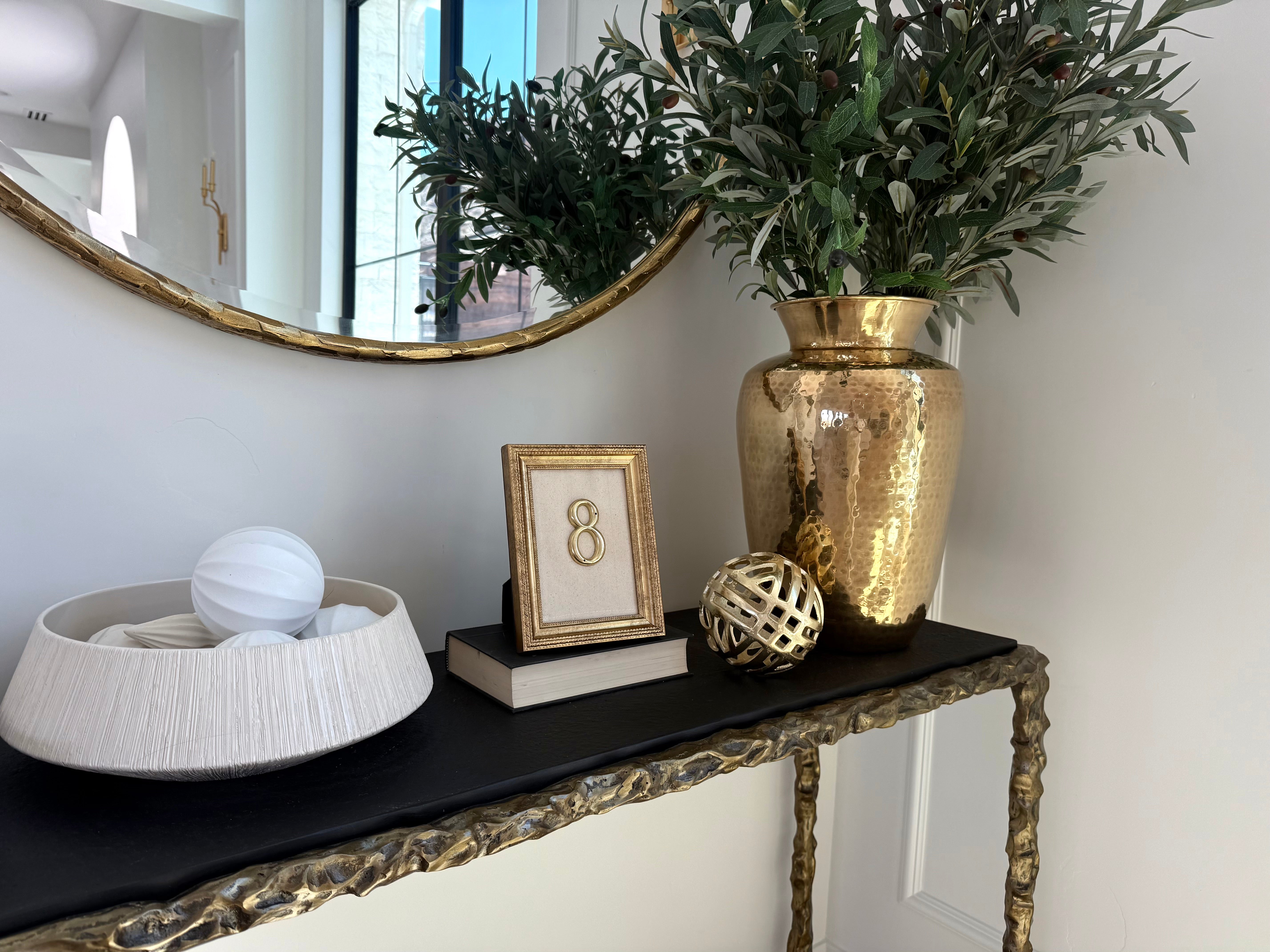 A styled console table corner with a large hammered gold vase of green foliage, a black tabletop, a gold-framed picture, a white textured bowl, and decorative gold and white spheres against a white wall.